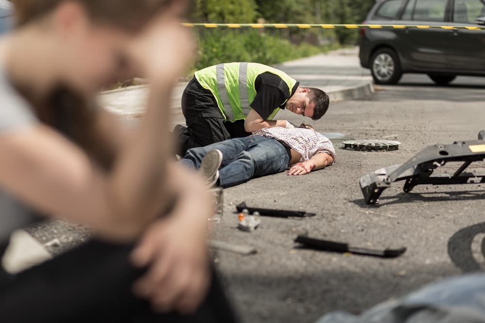 A paramedic assists an injured man on the street after a car accident, with a distressed woman sitting in the foreground.