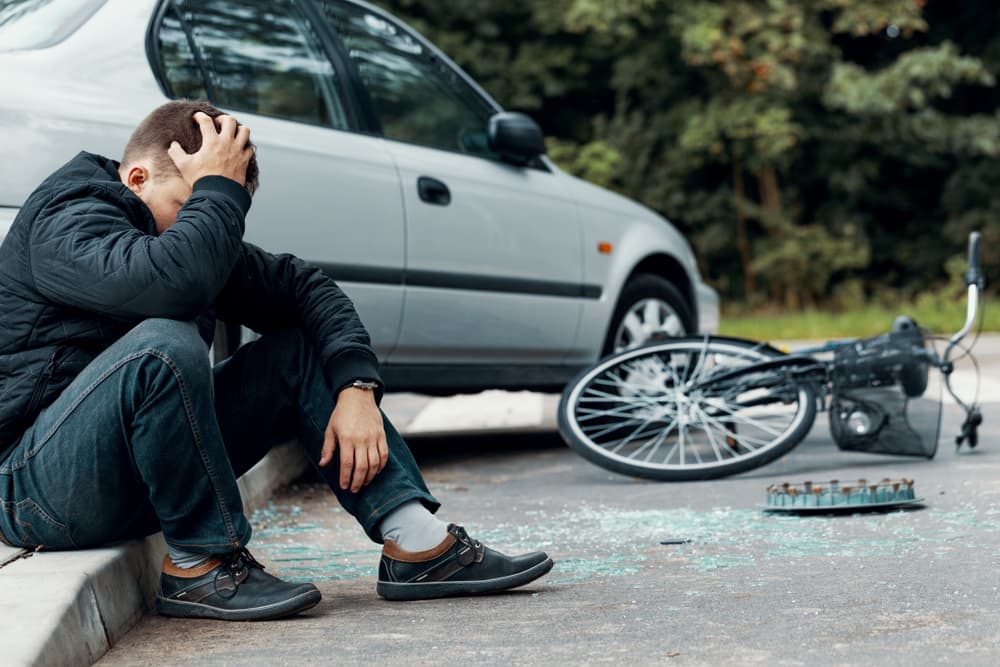 Worried biker holding his head suffered concussion and sitting on a pavement next to a car and bike crash